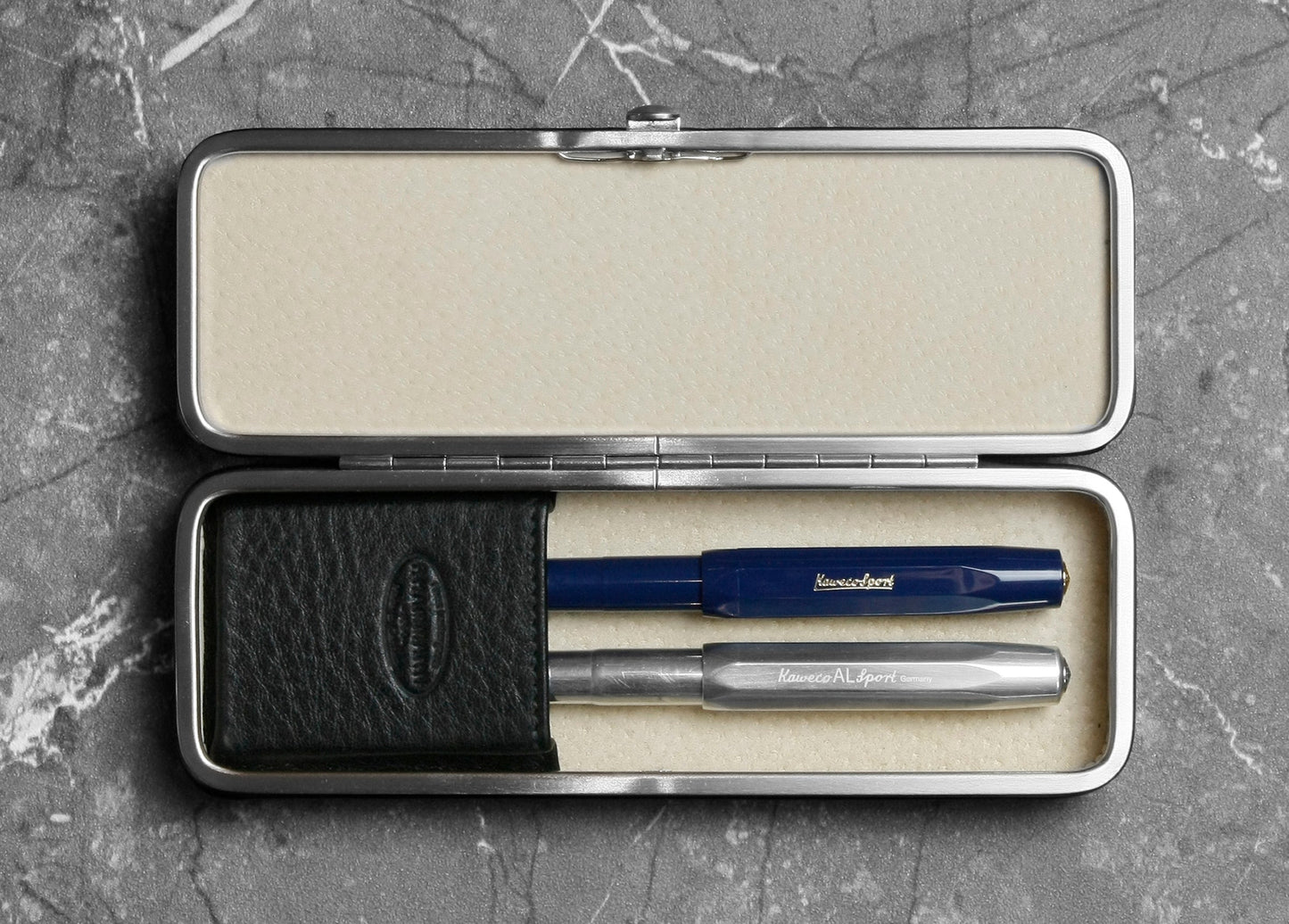 Open F. Hammann 3983 framed leather pen case holding two Kaweco fountain pens securely under its hidden flexible loops on a marble background.